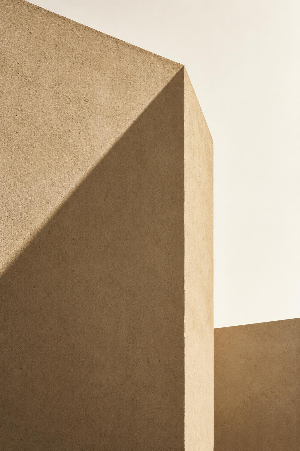 A sharp sand-colored concrete wall edge against bright sky, crossed by one crisp diagonal shadow